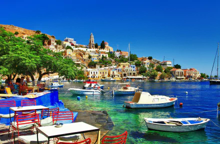 A serene view of Halki, Greece, featuring colorful boats anchored in clear waters, with charming man-made buildings lining the vibrant coastline under a bright blue sky.