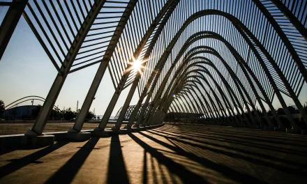 Sunset at a Greek Olympic Park: dramatic repeating steel arches casting long shadows, sunburst through ribs — 5K Ultra HD photography desktop wallpaper/background.