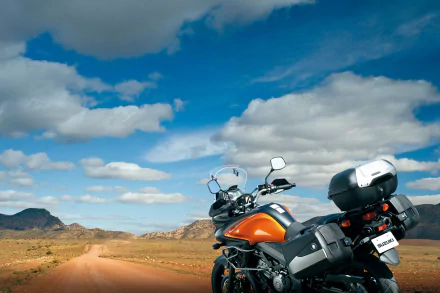 Orange Suzuki V-Strom motorcycle (bike/vehicle) parked on a desert dirt road under a blue sky with scattered clouds — HD PC desktop wallpaper and background.