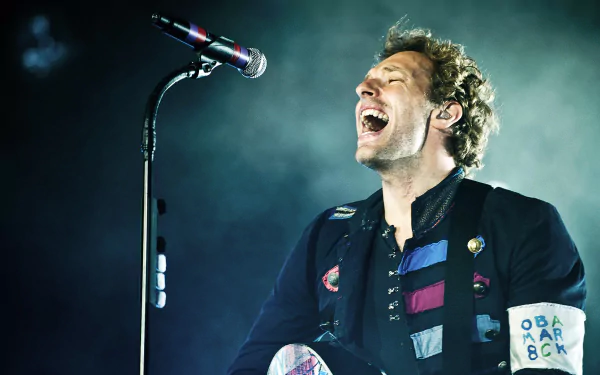 A dynamic HD wallpaper featuring a passionate Coldplay performance, showcasing the lead singer singing joyfully into a microphone under dramatic lighting.