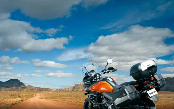 Orange Suzuki V-Strom motorcycle (bike/vehicle) parked on a desert dirt road under a blue sky with scattered clouds — HD PC desktop wallpaper and background.