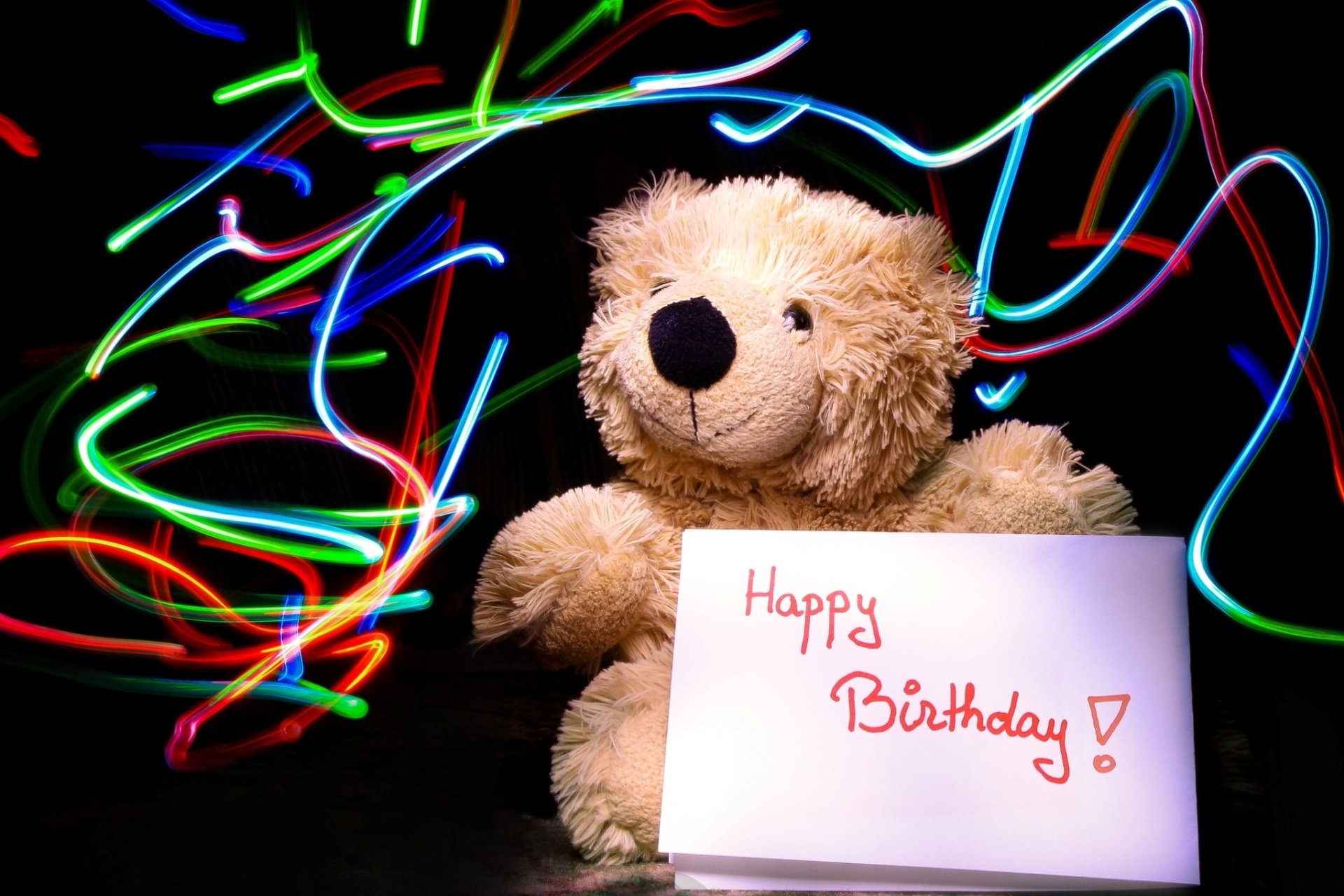 HD desktop wallpaper featuring a stuffed teddy bear holding a Happy Birthday! sign, surrounded by colorful light trails, creating a festive holiday birthday atmosphere.