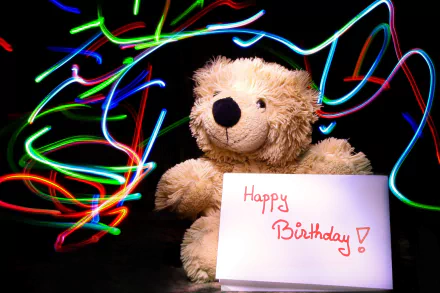 HD desktop wallpaper featuring a stuffed teddy bear holding a Happy Birthday! sign, surrounded by colorful light trails, creating a festive holiday birthday atmosphere.
