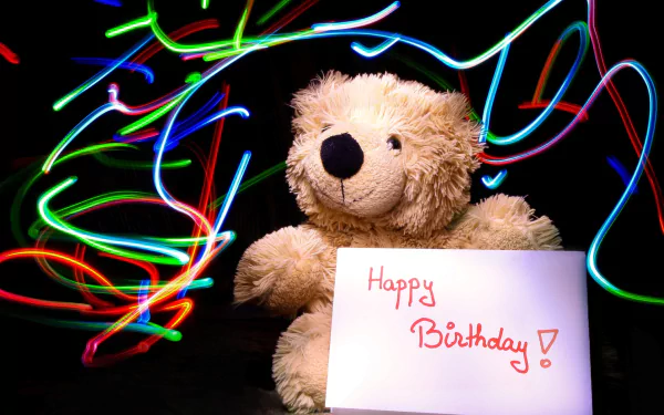 HD desktop wallpaper featuring a stuffed teddy bear holding a Happy Birthday! sign, surrounded by colorful light trails, creating a festive holiday birthday atmosphere.