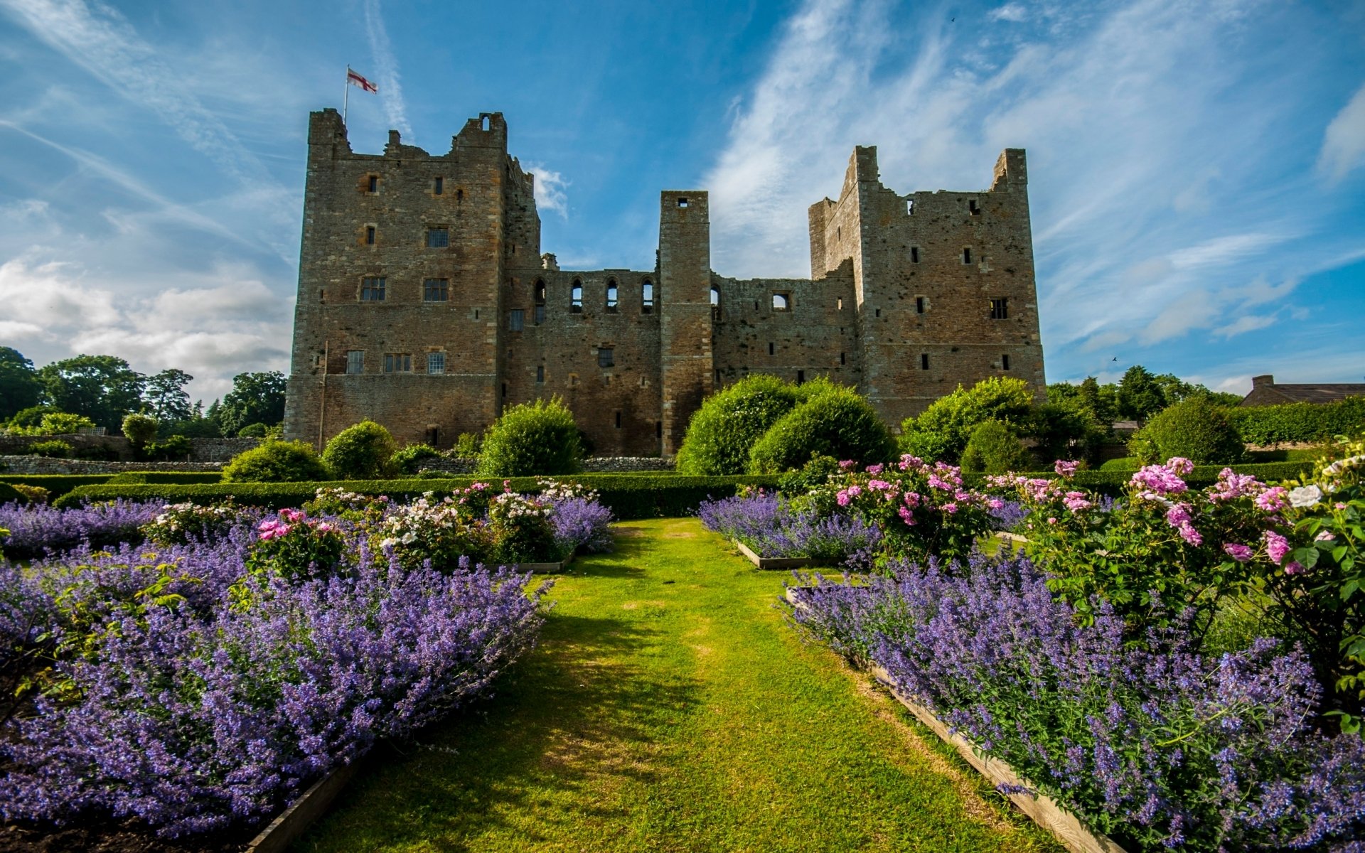 A scenic view of Bolton Castle, showcasing its historic structure framed by vibrant gardens of blooming roses and purple flowers against a clear blue sky.