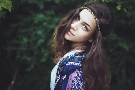 Moody portrait of a young woman with loose waves and a beaded headband, gazing over her shoulder against blurred green foliage — HD PC desktop wallpaper/background.