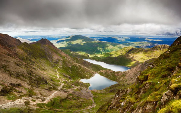 A stunning 4K Ultra HD landscape of Snowdonia, Wales, featuring misty clouds above hills, a serene lake, and rugged mountains under a dramatic sky.