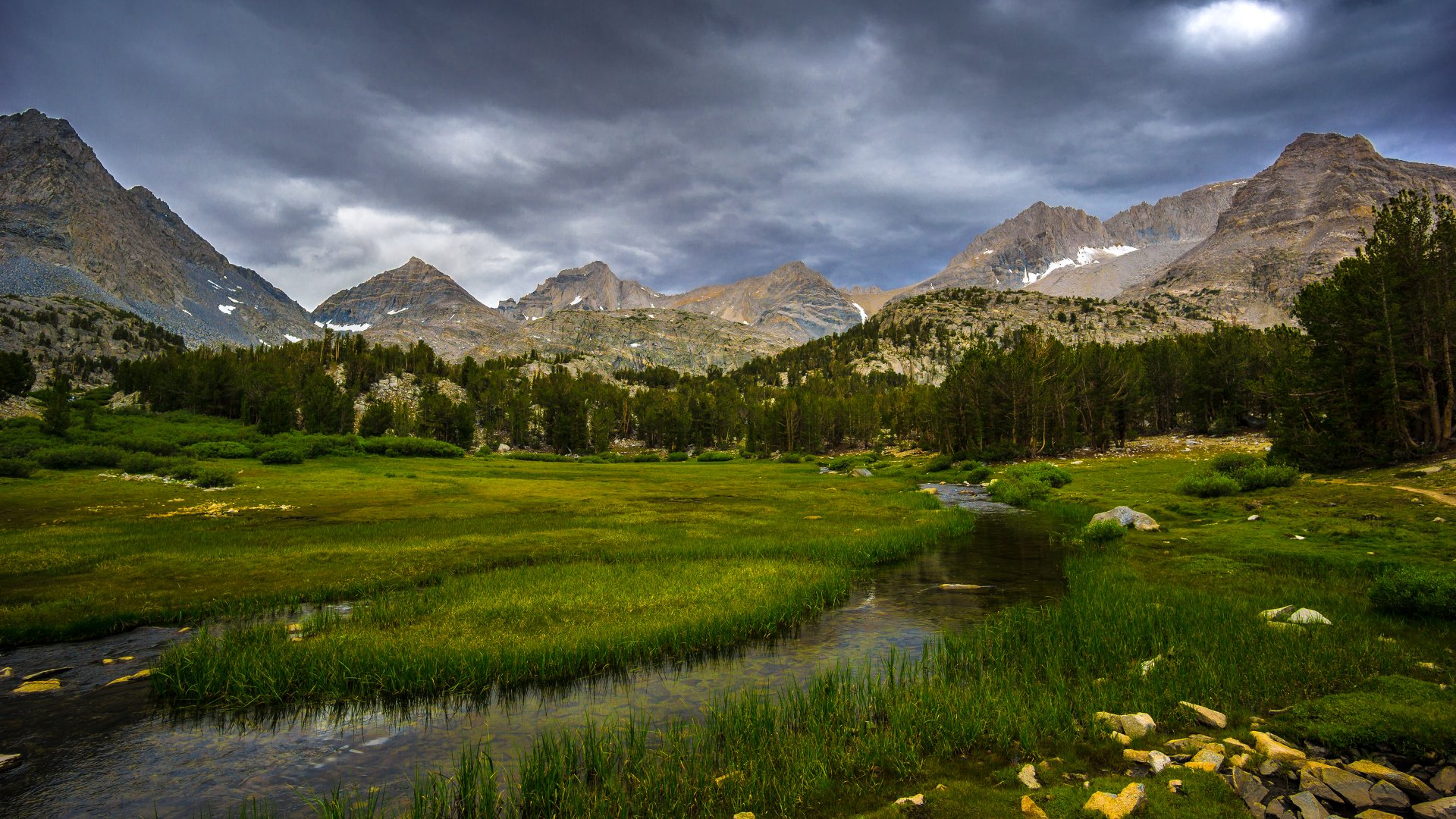 A serene landscape featuring lush green meadows and a flowing stream, framed by majestic mountains under a dramatic, cloudy sky—an inspiring HD nature wallpaper.