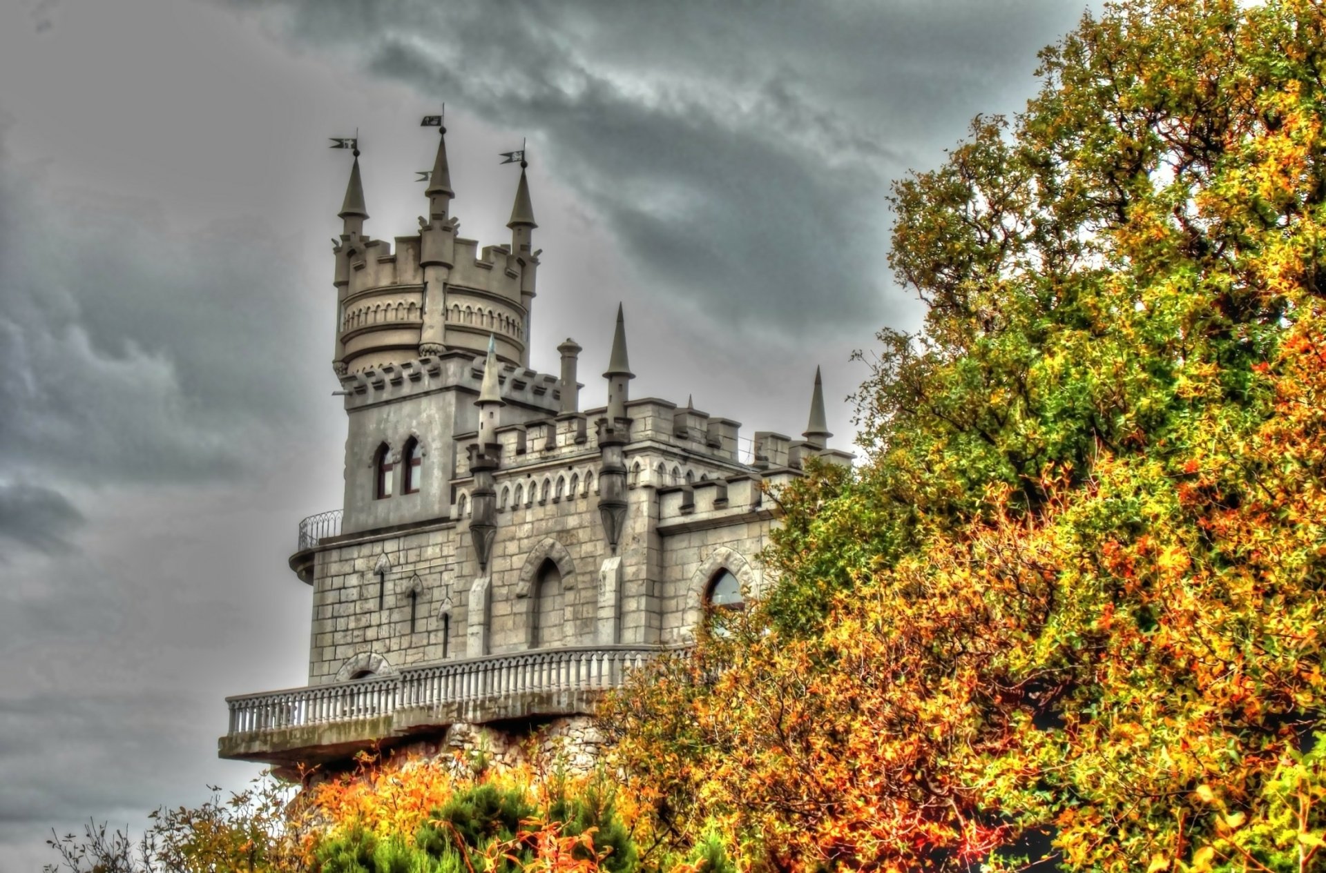 HD wallpaper of Russia's iconic Swallow's Nest castle perched above colorful autumn foliage in Yalta, showcasing intricate man-made architecture against a cloudy sky.