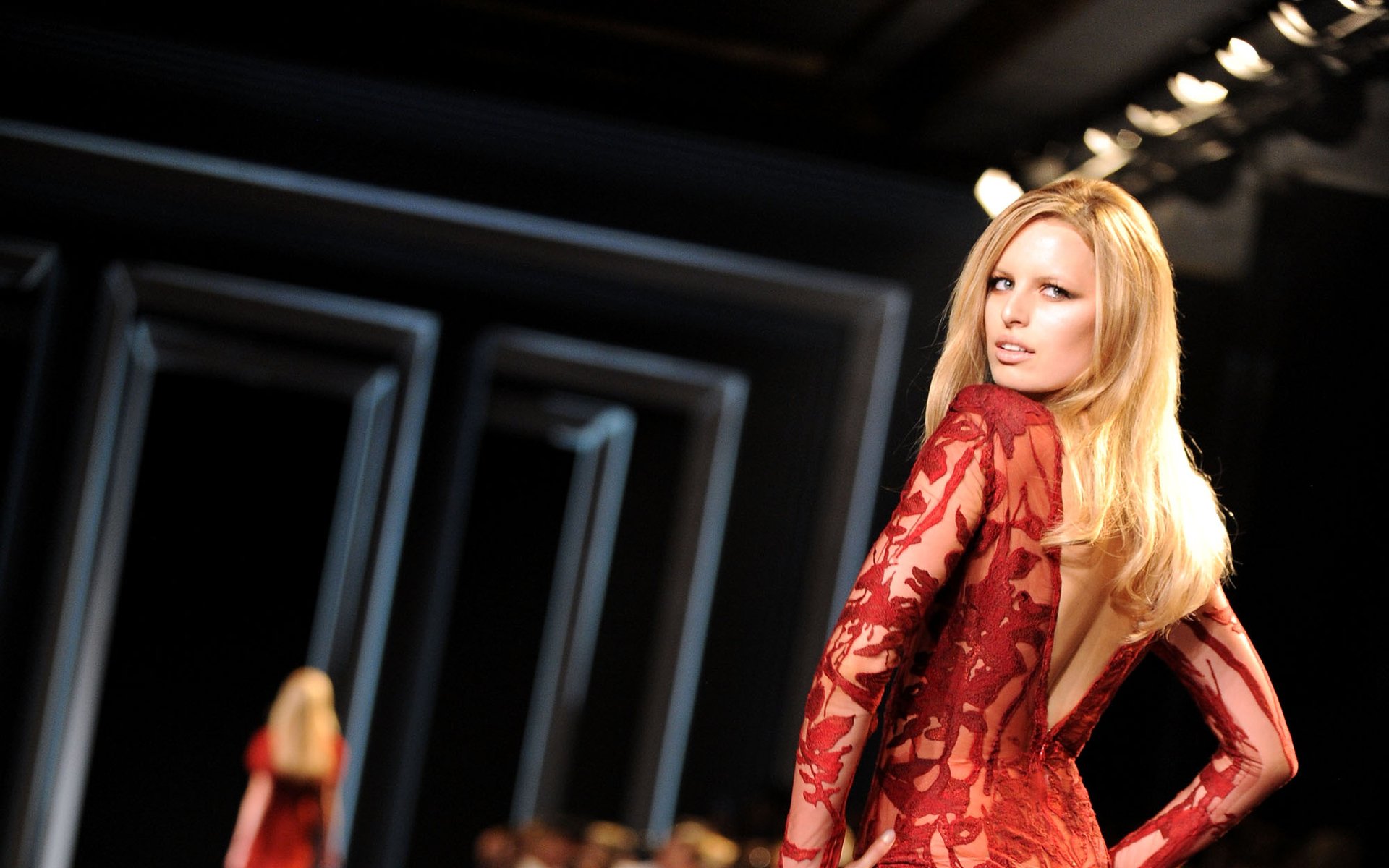 HD desktop wallpaper of a blonde runway model in a red lace, backless gown looking over her shoulder under dramatic stage lights.