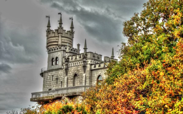 HD wallpaper of Russia's iconic Swallow's Nest castle perched above colorful autumn foliage in Yalta, showcasing intricate man-made architecture against a cloudy sky.