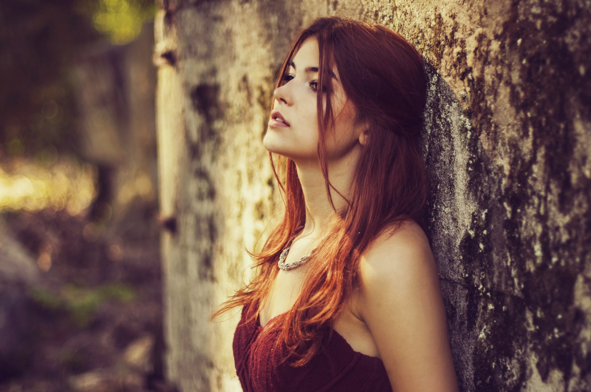 A woman with red hair leans against a textured wall, her eyes closed, exuding a contemplative mood. This HD desktop wallpaper and background captures a serene moment in nature.