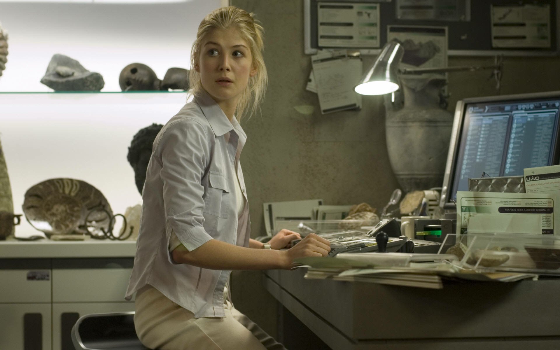 HD PC desktop wallpaper showing a scene from the movie Doom: a woman in a lab shirt at a cluttered desk, glancing back toward the camera amid monitors and specimen displays.