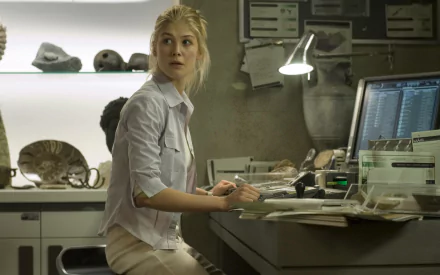 HD PC desktop wallpaper showing a scene from the movie Doom: a woman in a lab shirt at a cluttered desk, glancing back toward the camera amid monitors and specimen displays.