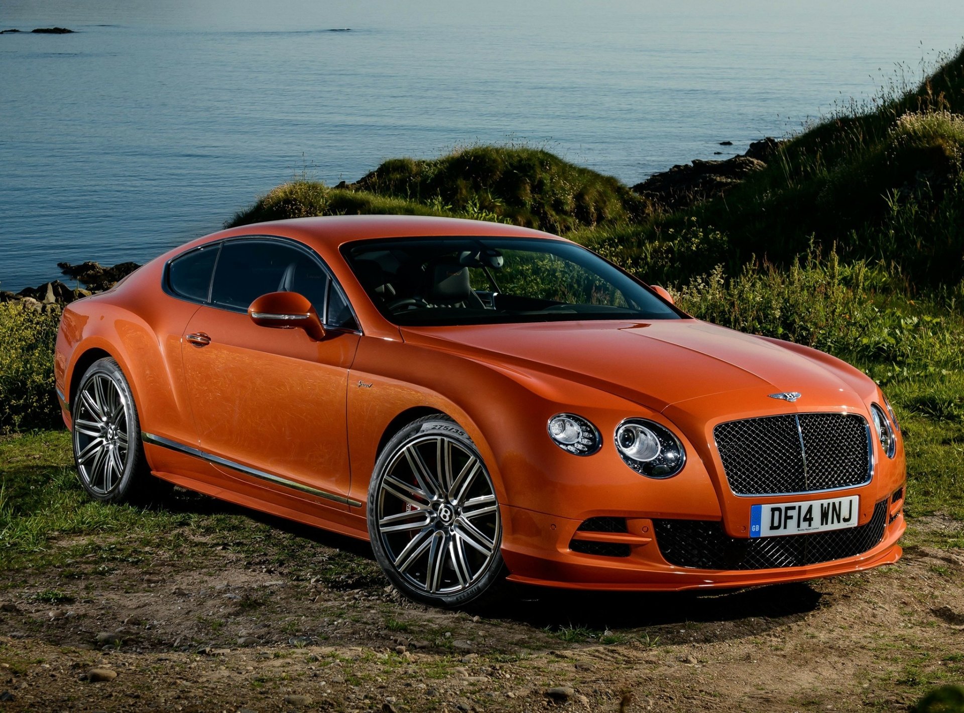 HD desktop wallpaper showing an orange Bentley Continental parked on a grassy area near the water with hills in the background.