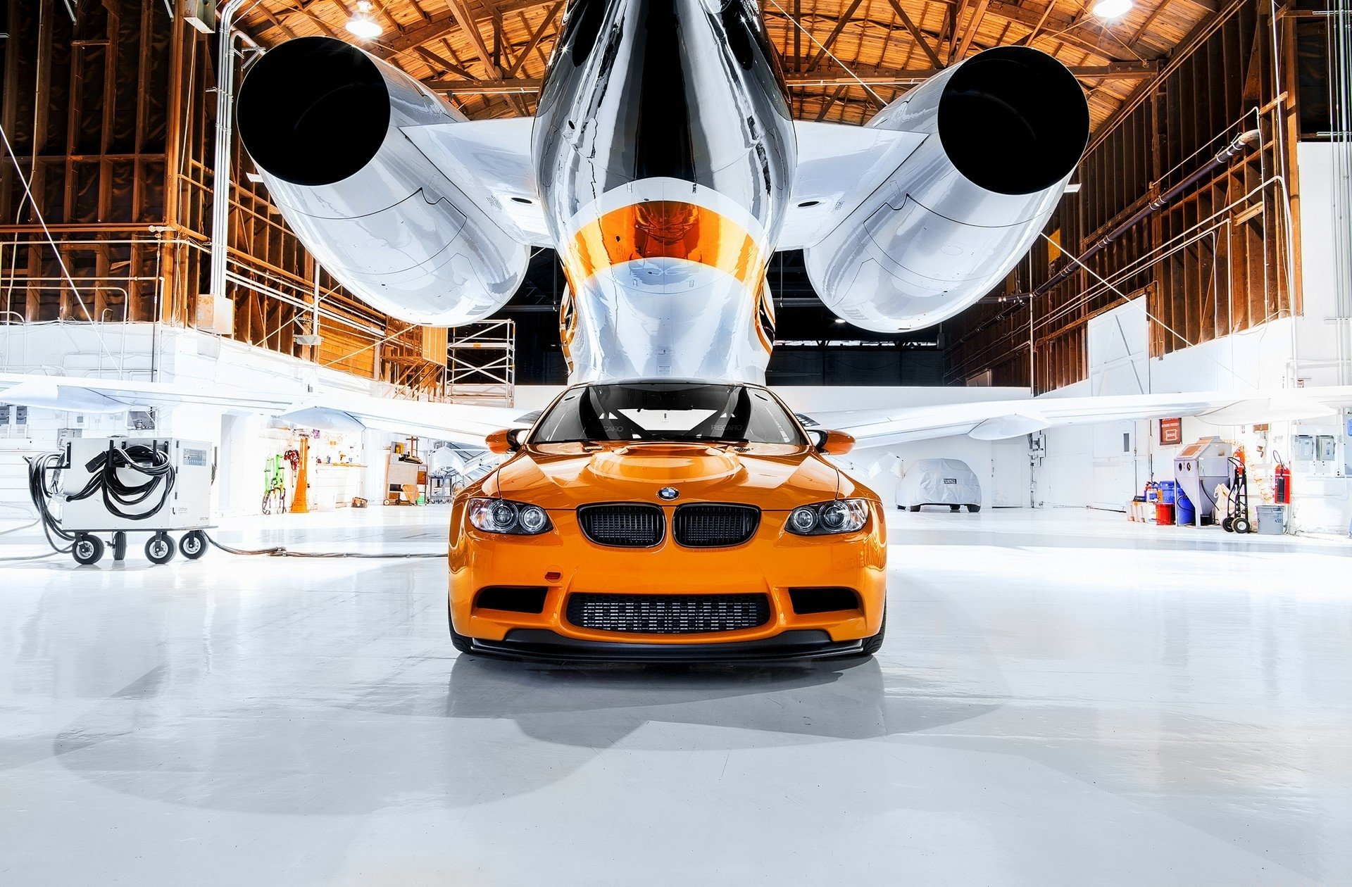 HD PC desktop wallpaper featuring an orange BMW vehicle prominently displayed inside a bright, industrial-style hangar with sleek aircraft engines overhead.