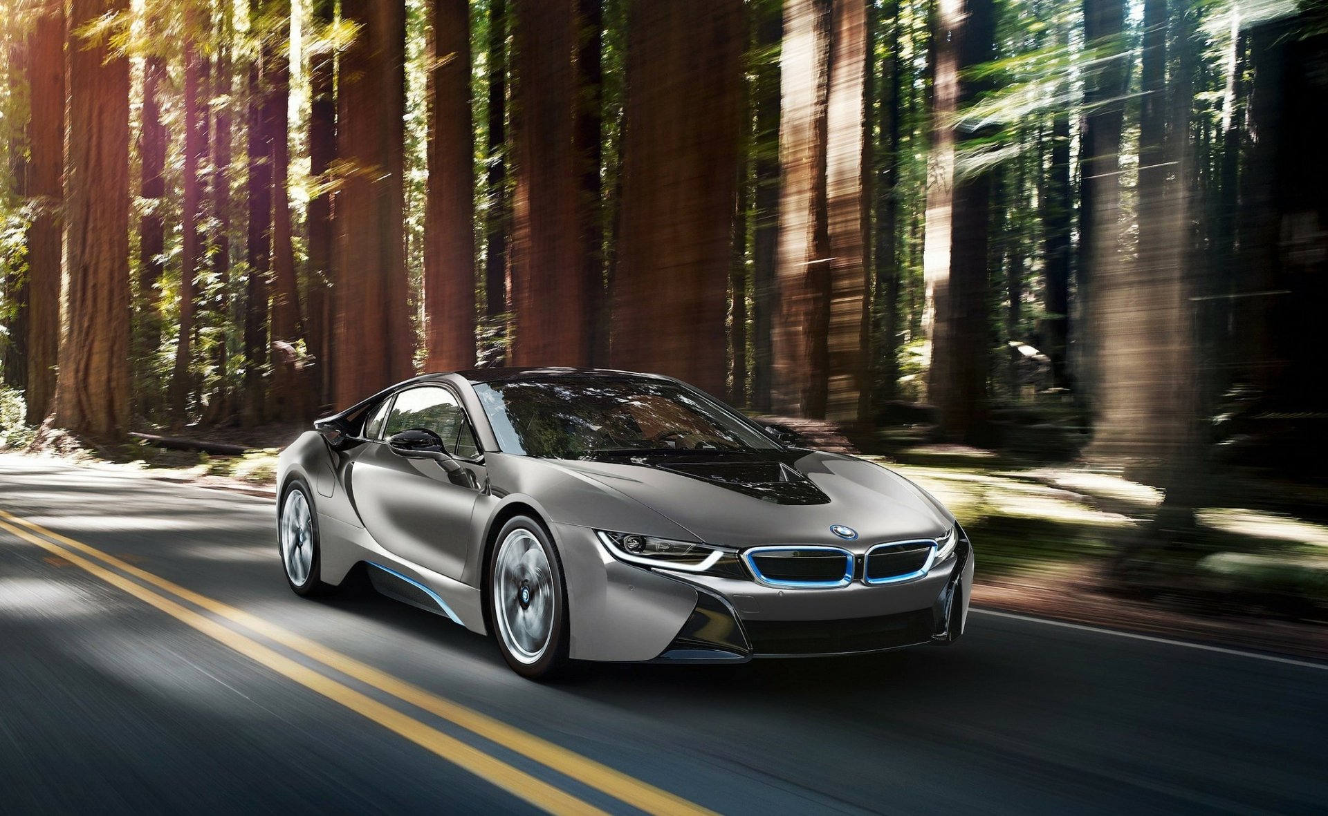 A silver BMW i8 sports car drives along a two-lane road surrounded by tall forest trees, captured in HD for a PC desktop wallpaper.