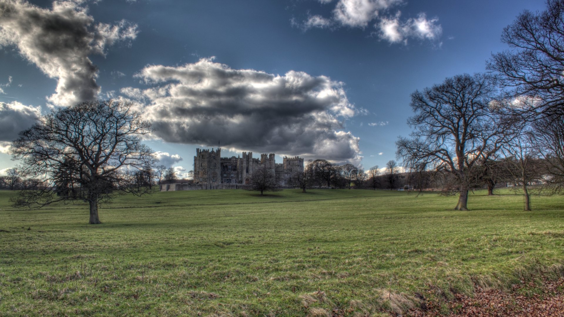 HD PC desktop wallpaper featuring the man-made structure of Raby Castle under a dramatic sky, framed by trees on a vast green lawn.