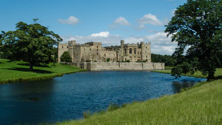 2K Quad HD PC desktop wallpaper: Raby Castle, a man-made stone fortress beside a reflective moat, set among green lawns and trees under a bright blue sky.