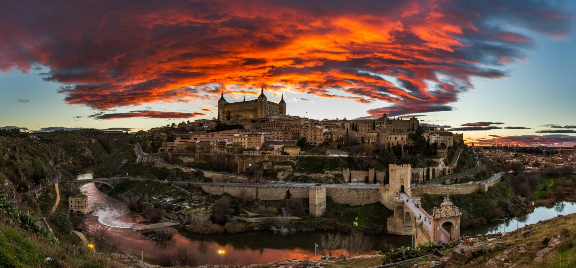 Sunset over the historic man-made cityscape of Toledo, Spain, with vibrant orange clouds illuminating the skyline in this HD desktop wallpaper.