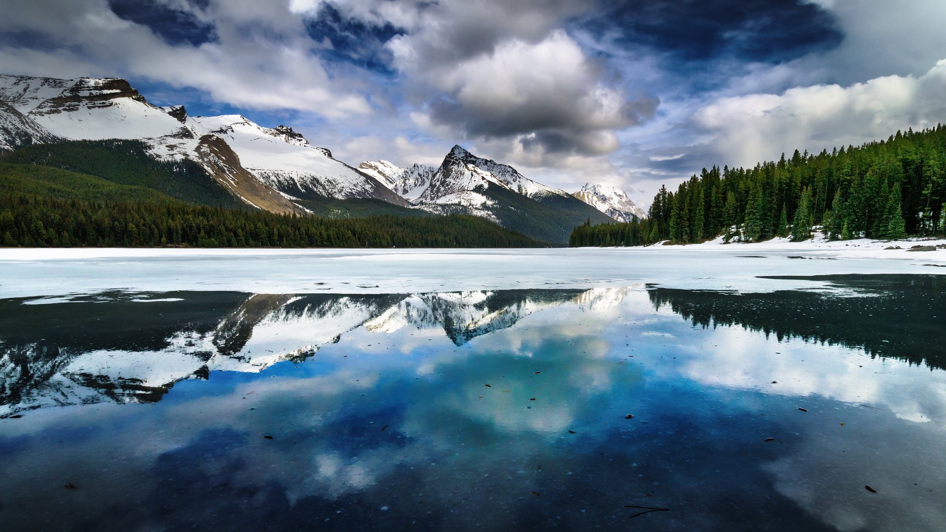 4K Ultra HD desktop wallpaper featuring snow-capped mountains and dense forest reflected vividly in a calm lake under a dramatic cloudy sky.