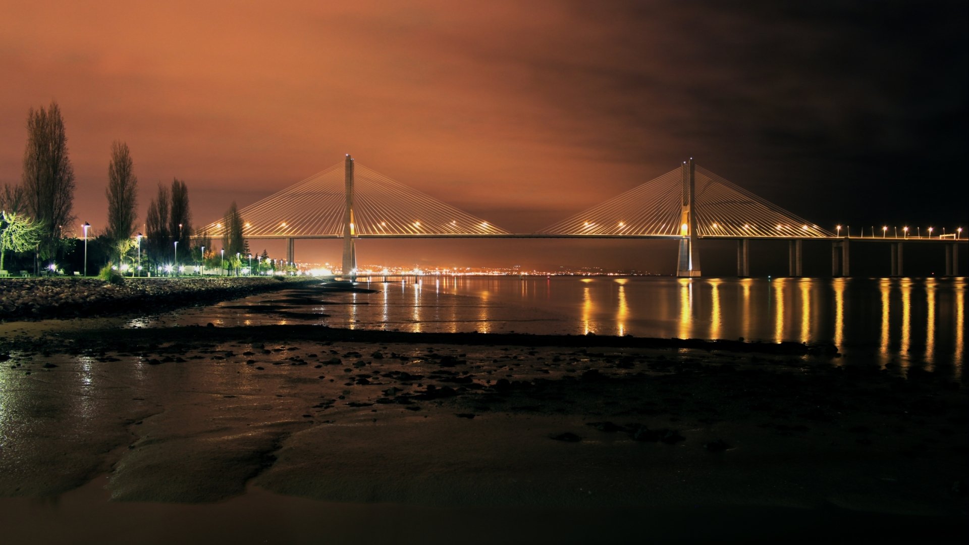 2K Quad HD desktop wallpaper: illuminated Vasco da Gama Bridge at night, golden lights reflected on calm river under a warm orange sky, shoreline in foreground.
