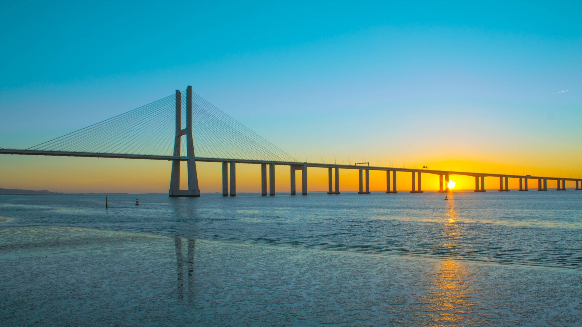 Man-made Vasco da Gama Bridge at sunrise spanning calm water with vivid sky and reflections — 2K Quad HD PC desktop wallpaper background.