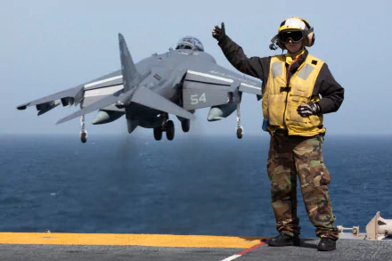 A McDonnell Douglas AV-8B Harrier II aircraft hovers above an amphibious assault ship with a navy Marine signaling on the flight deck.