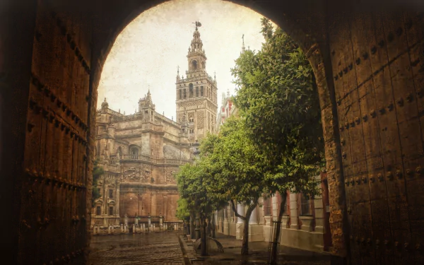 View of a historic street in Seville, Spain, framed by an arched doorway with a prominent man-made tower and lush trees, captured in HD for desktop wallpaper.