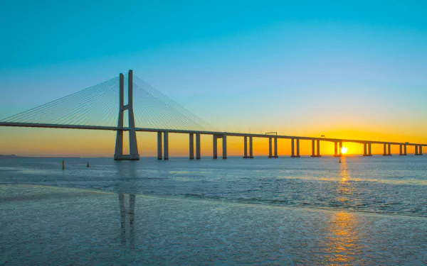 Man-made Vasco da Gama Bridge at sunrise spanning calm water with vivid sky and reflections — 2K Quad HD PC desktop wallpaper background.