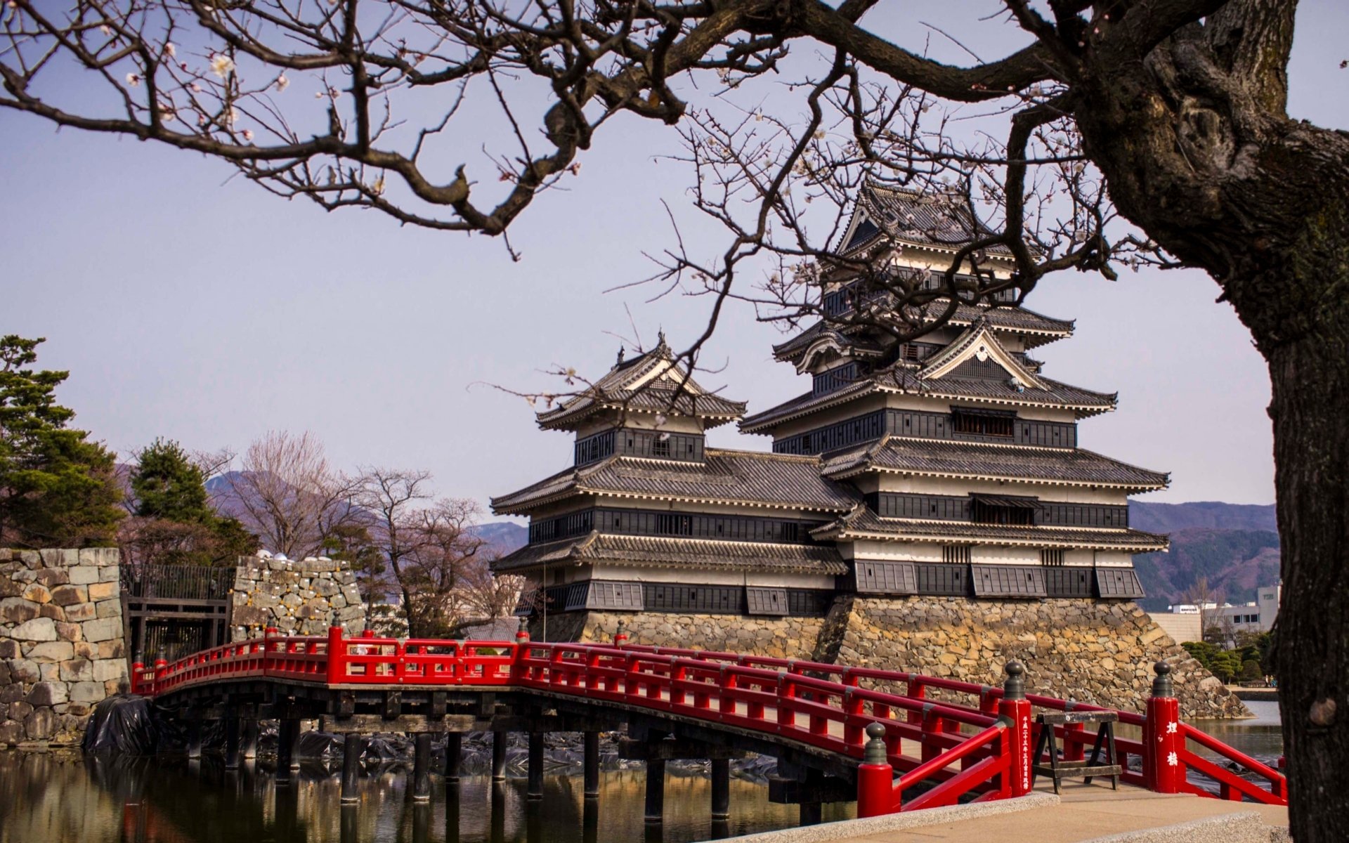 HD PC desktop wallpaper/background: man-made Matsumoto Castle — layered black keeps beside a red arched bridge over a reflective moat, framed by bare trees and a pale winter sky.