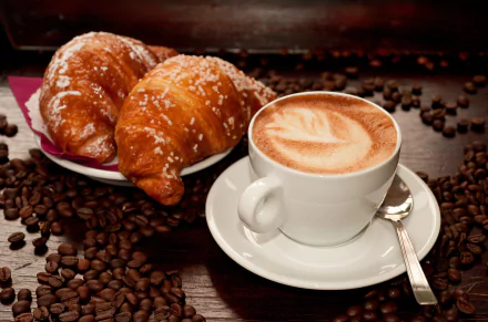 A warm cappuccino sits beside golden croissants on a simple plate, surrounded by scattered coffee beans, creating a cozy, inviting scene in this 8K Ultra HD desktop wallpaper.