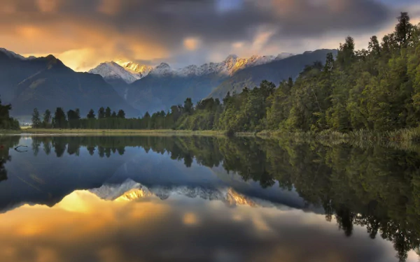 Aoraki/Mount Cook in New Zealand reflected in a calm lake, surrounded by lush woodlands under a cloudy sky at sunset.