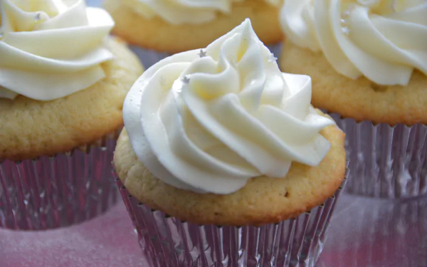 Close-up 4K Ultra HD PC desktop wallpaper of vanilla cupcakes with swirled white frosting in silver liners on a pink surface.