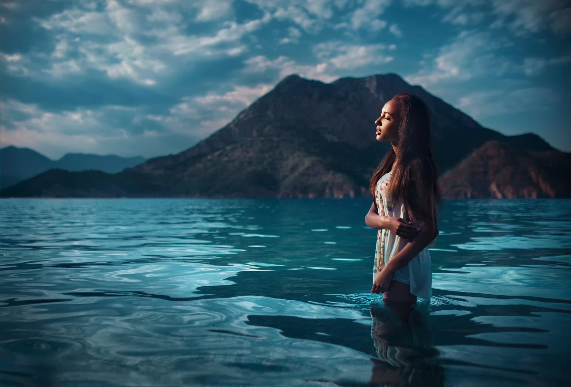 A woman stands in serene blue waters with a mountainous backdrop in Turkey, creating a captivating scene. This HD desktop wallpaper and background image features a model named Mary Senn.