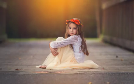 A little girl sits on a pathway, wearing a white sweater and a light skirt, with a flower crown in her hair. The scene is illuminated by warm, glowing light, creating a captivating moment.