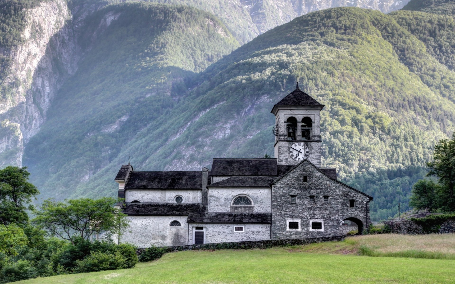 HD PC desktop wallpaper featuring a serene stone church nestled in lush green hills with towering mountains in the background, evoking a peaceful religious atmosphere.