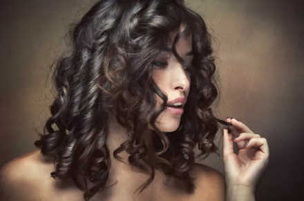 4K Ultra HD wallpaper of a woman with dark, curly hair softly holding a curl, her face partially turned to the side against a muted brown background.
