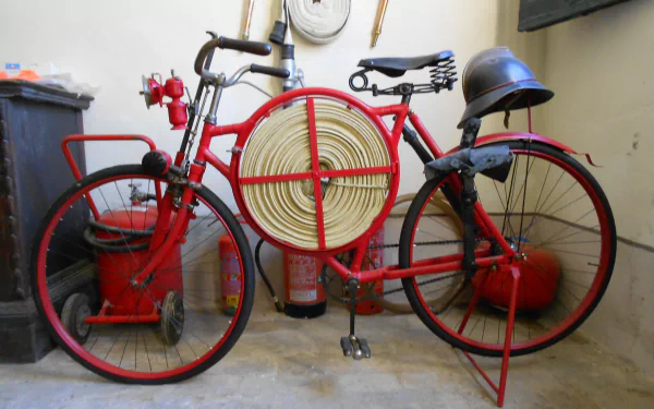 A vintage red bicycle with a fire hose reel and helmet, set against a backdrop of fire extinguishers. This 4K Ultra HD wallpaper captures a unique vehicle scene.