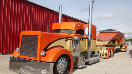 HD desktop wallpaper featuring a vibrant orange Peterbilt truck and trailer parked beside a red building under a partly cloudy sky.