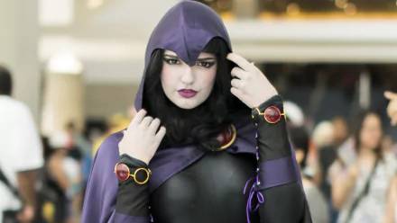 A woman in a Raven (DC Comics) cosplay poses confidently, wearing a dark hooded outfit and accessories, set against a vibrant convention backdrop. High-definition desktop wallpaper.