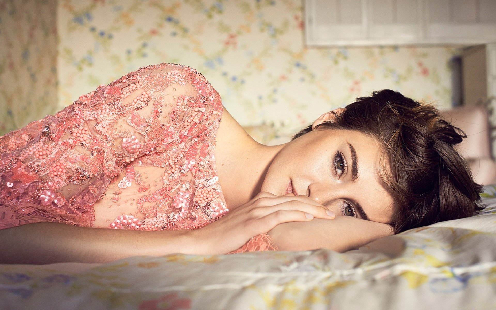 Brunette American actress Shailene Woodley lies on a bed wearing a pink lace top, featured in an HD PC desktop wallpaper and background.