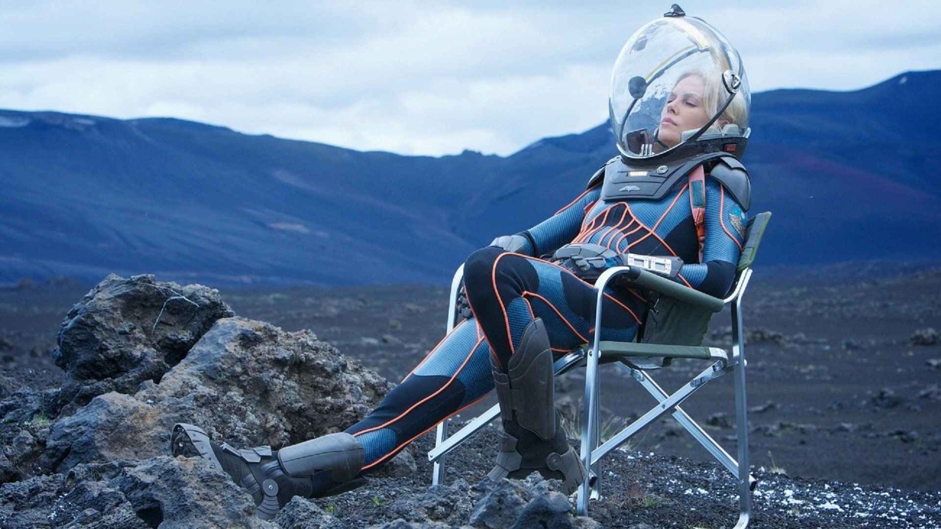 Charlize Theron in a spacesuit from the movie Prometheus sits relaxed on a chair against a rocky, mountainous landscape in this HD PC desktop wallpaper.