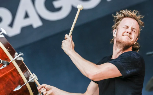 A dynamic performance by an American alternative band, Imagine Dragons, captures the energy of a drummer on stage, set against their iconic backdrop in vibrant detail.