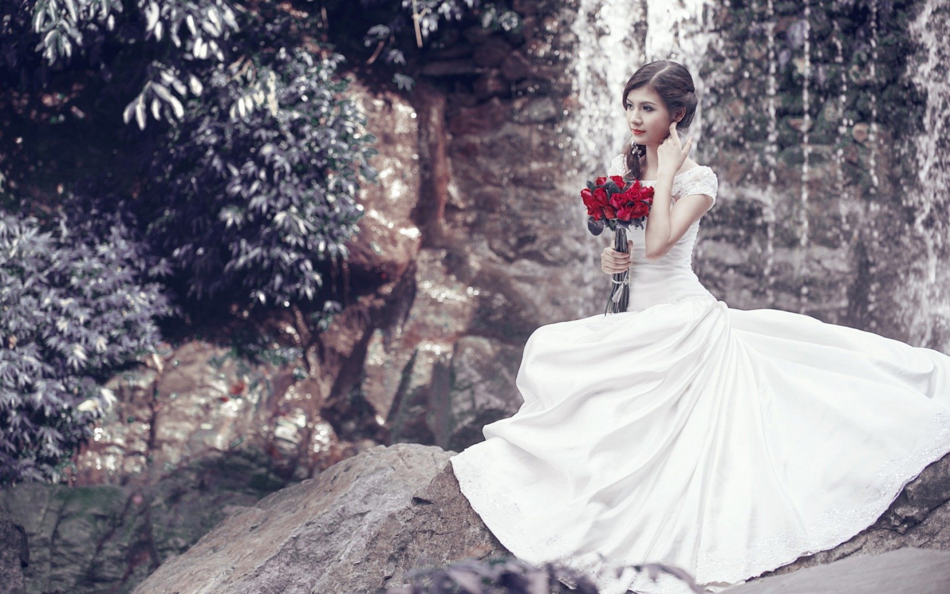 HD PC desktop wallpaper of an Asian woman in a flowing white dress holding a bouquet of red roses, seated on a rock with a waterfall and lush foliage in the background.