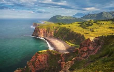 HD desktop wallpaper of Kamchatka's rugged coastline in Russia's Far East, featuring green cliffs, a sandy beach, and a vast ocean under a partly cloudy sky.