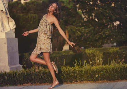 A woman in a floral dress joyfully walks outdoors, holding a bag, surrounded by greenery. The image features Clara Alonso and serves as a vibrant HD desktop wallpaper.