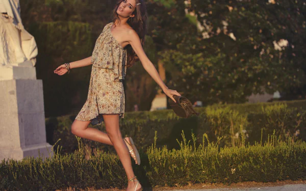 A woman in a floral dress joyfully walks outdoors, holding a bag, surrounded by greenery. The image features Clara Alonso and serves as a vibrant HD desktop wallpaper.