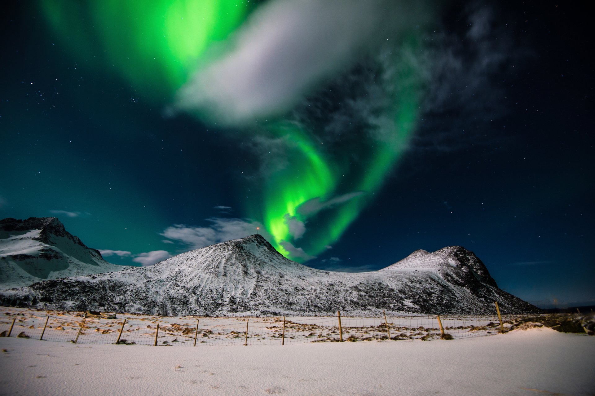 A stunning HD wallpaper featuring the Aurora Borealis illuminating the night sky over the snowy mountains of Lofoten Islands, Norway. The natural landscape is bathed in green aurora lights under a dark sky.