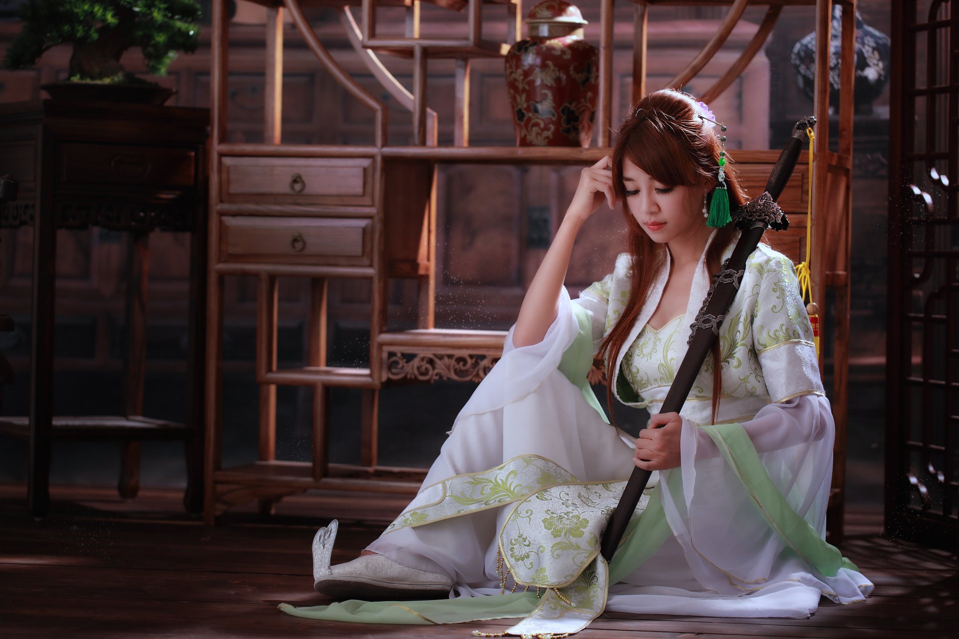 A woman in traditional Asian dress sits gracefully with a sword, embodying the essence of elegance and strength. The backdrop features traditional wooden decor. A stunning HD wallpaper.